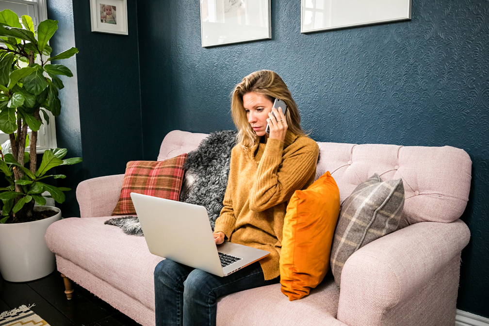 A woman sits on the sofa using her laptop while on the phone