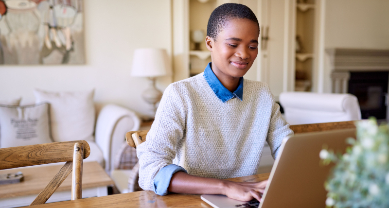 A lady sitting at a table, smiling, whilst using a laptop